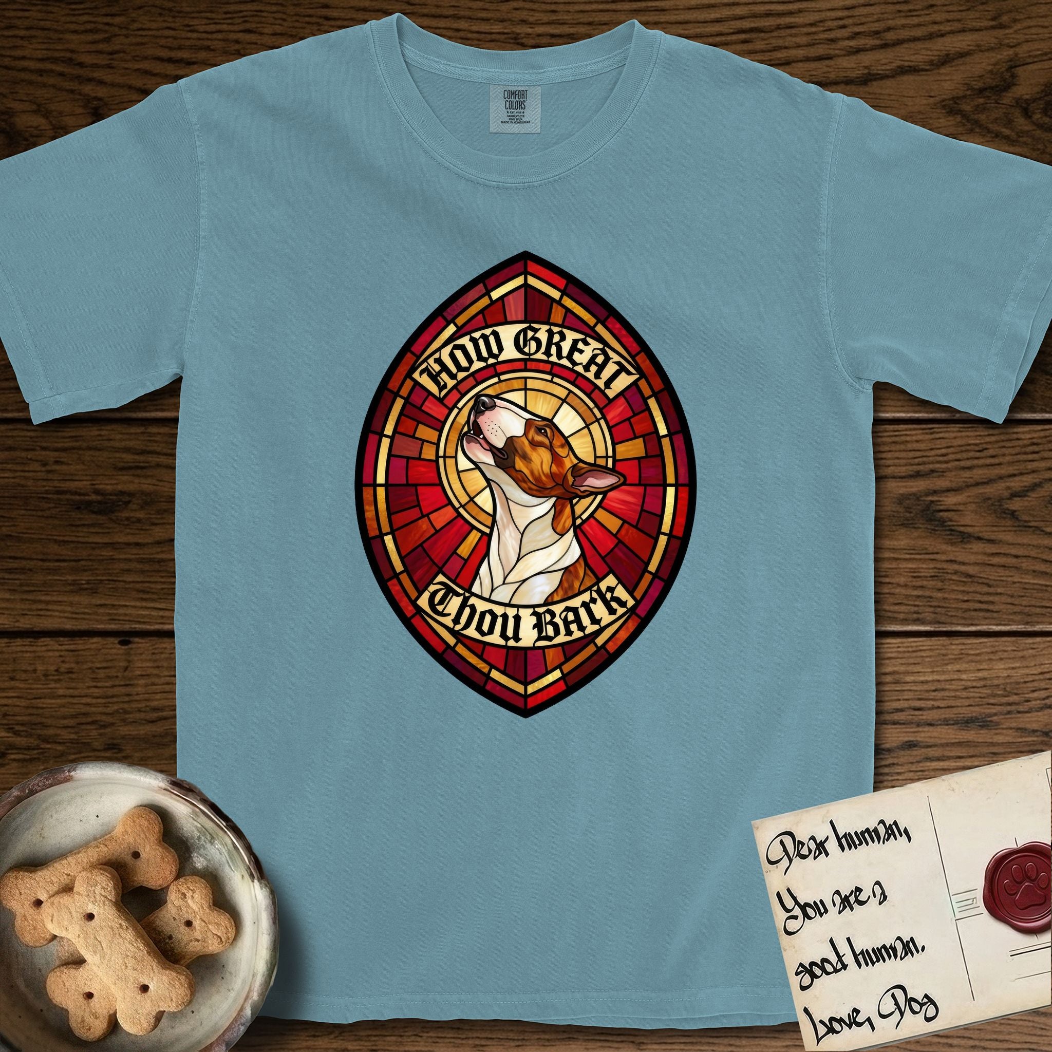How Great Thou Bark Bull Terrier T-Shirt flat lay in Ice Blue, featuring a full-color stained-glass saint window of a brown and white Bull Terrier with head raised and mouth open mid-howl, haloed in golden light.