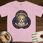 How Great Thou Bark Coton de Tulear T-Shirt flat lay in Blossom, featuring a full-color stained-glass saint window of a sable and white Coton de Tulear facing forward, haloed in golden light.