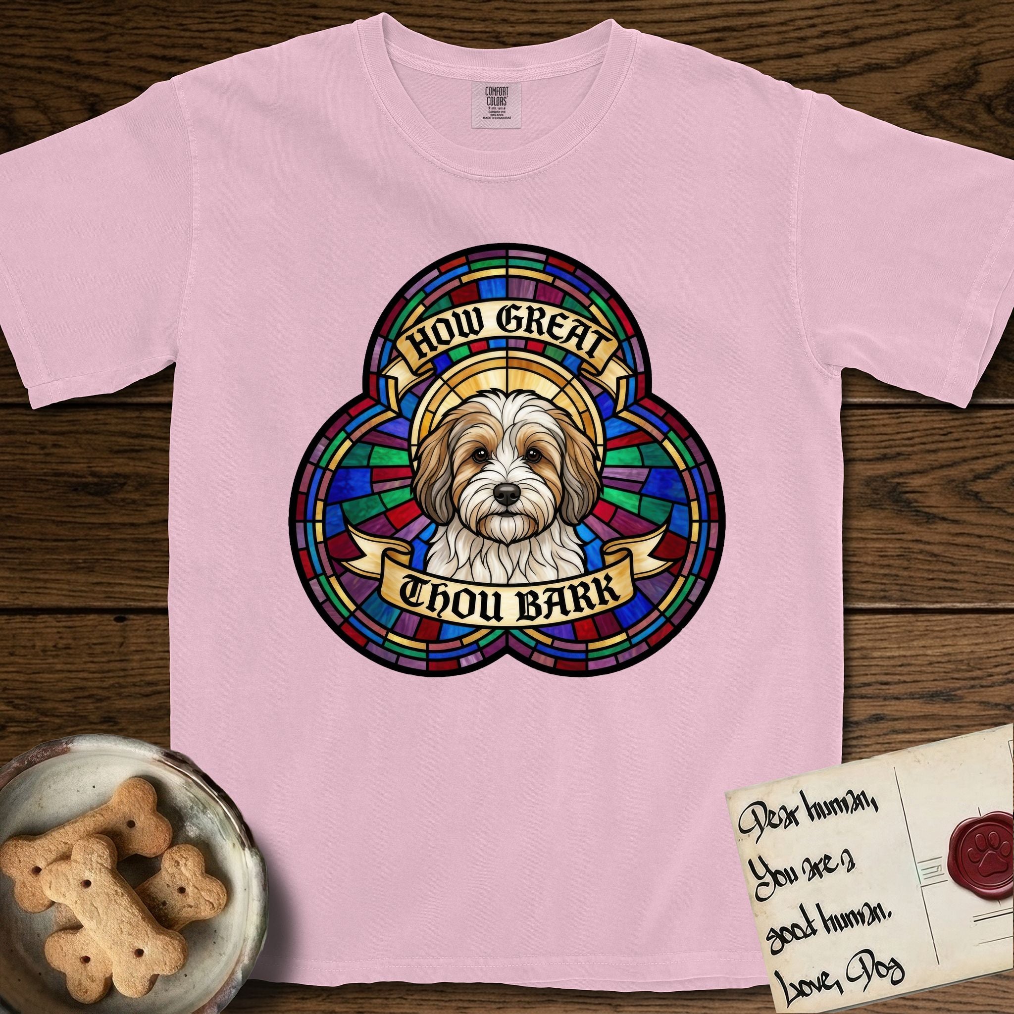 How Great Thou Bark Coton de Tulear T-Shirt flat lay in Blossom, featuring a full-color stained-glass saint window of a sable and white Coton de Tulear facing forward, haloed in golden light.