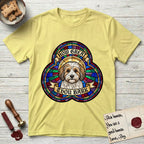 How Great Thou Bark Coton de Tulear T-Shirt flat lay in Cornsilk, featuring a full-color stained-glass saint window of a sable and white Coton de Tulear facing forward, haloed in golden light.