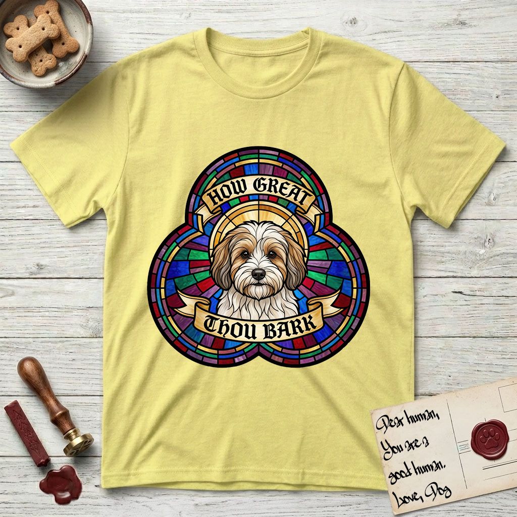 How Great Thou Bark Coton de Tulear T-Shirt flat lay in Cornsilk, featuring a full-color stained-glass saint window of a sable and white Coton de Tulear facing forward, haloed in golden light.