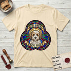 How Great Thou Bark Coton de Tulear T-Shirt flat lay in Natural, featuring a full-color stained-glass saint window of a sable and white Coton de Tulear facing forward, haloed in golden light.