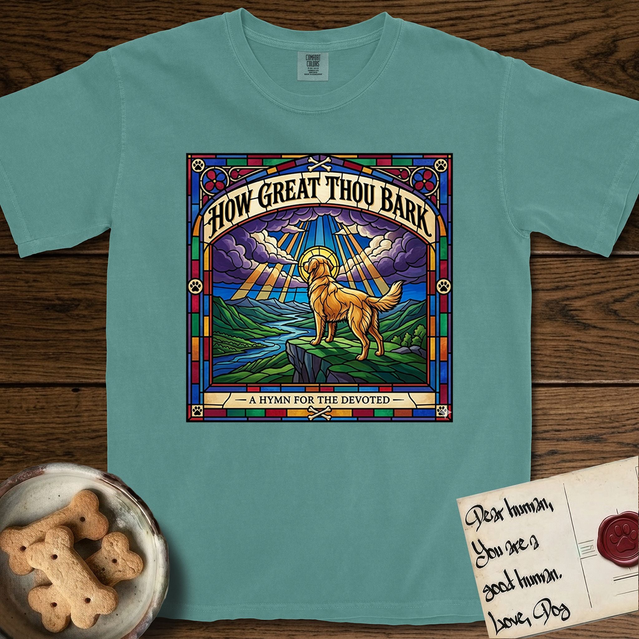 How Great Thou Bark Golden Retriever T-Shirt flat lay in Blue Spruce, featuring a full-color stained-glass saint window of a Golden Retriever standing on a mountain with light breaking through the clouds and a halo.