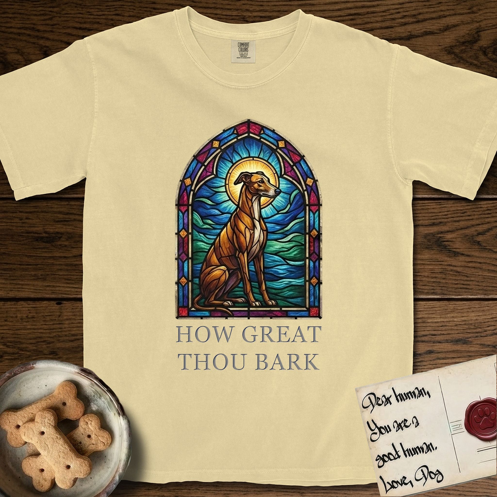 How Great Thou Bark Greyhound T-Shirt flat lay in Butter, featuring a full-color stained-glass saint window of a Greyhound sitting in stillness, with a halo.
