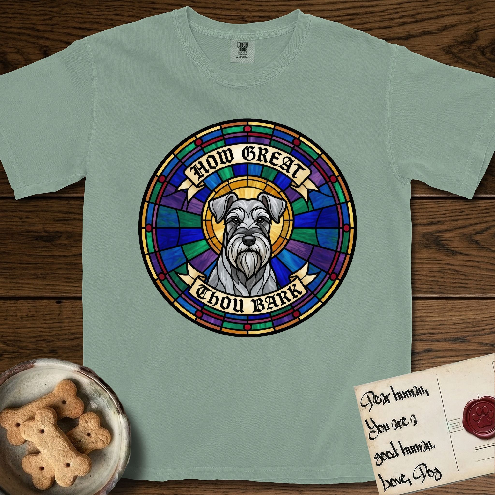 How Great Thou Bark Schnauzer T-Shirt flat lay in Bay, featuring a full-color stained-glass saint window of a salt-and-pepper Schnauzer with bushy eyebrows and beard, haloed in golden light.