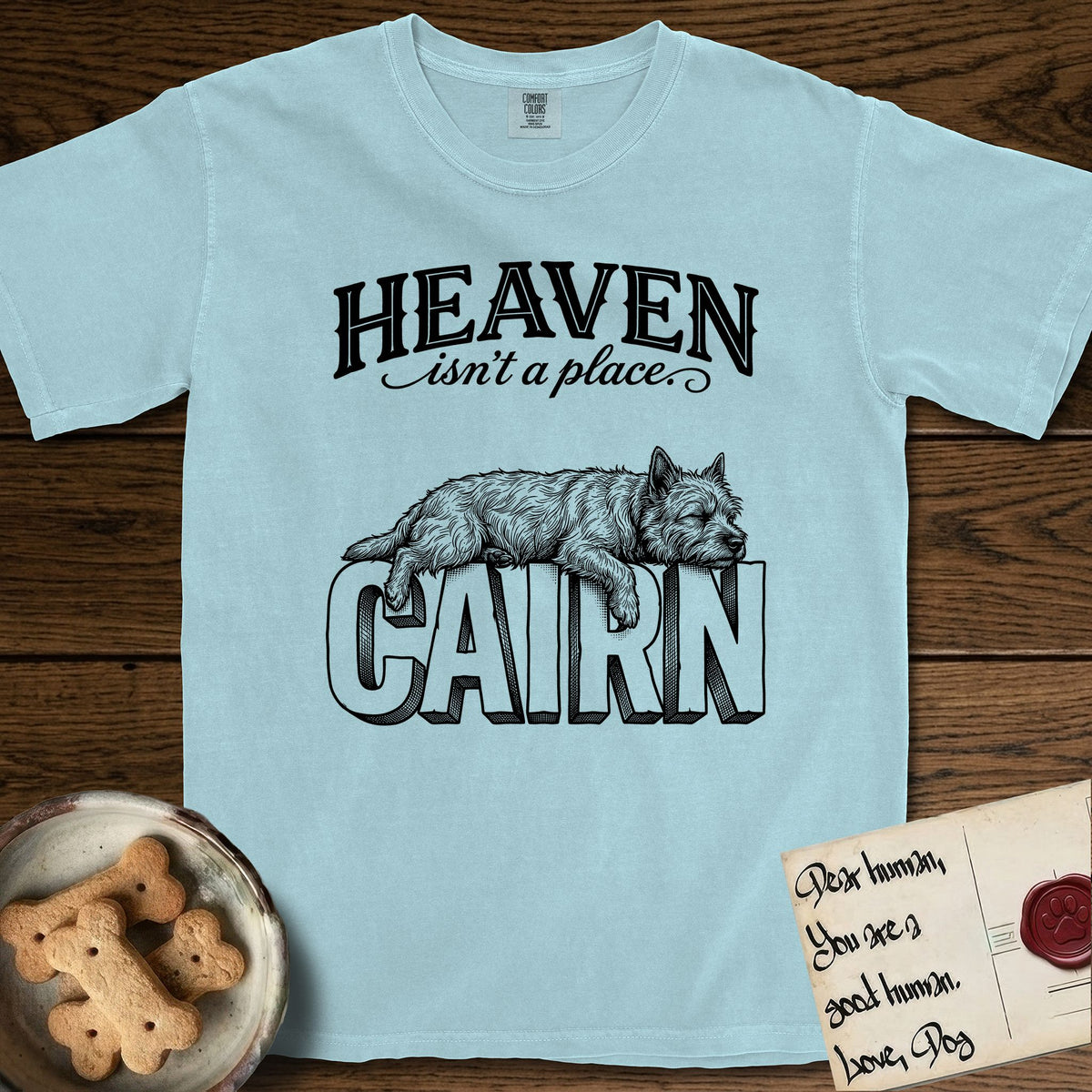 Heaven Isn't a Place Cairn Terrier T-Shirt flat lay in Chambray, featuring Black ink engraving illustration of a shaggy Cairn Terrier asleep on top of the giant carved block letters CAIRN, under the lettering HEAVEN isn't a place..
