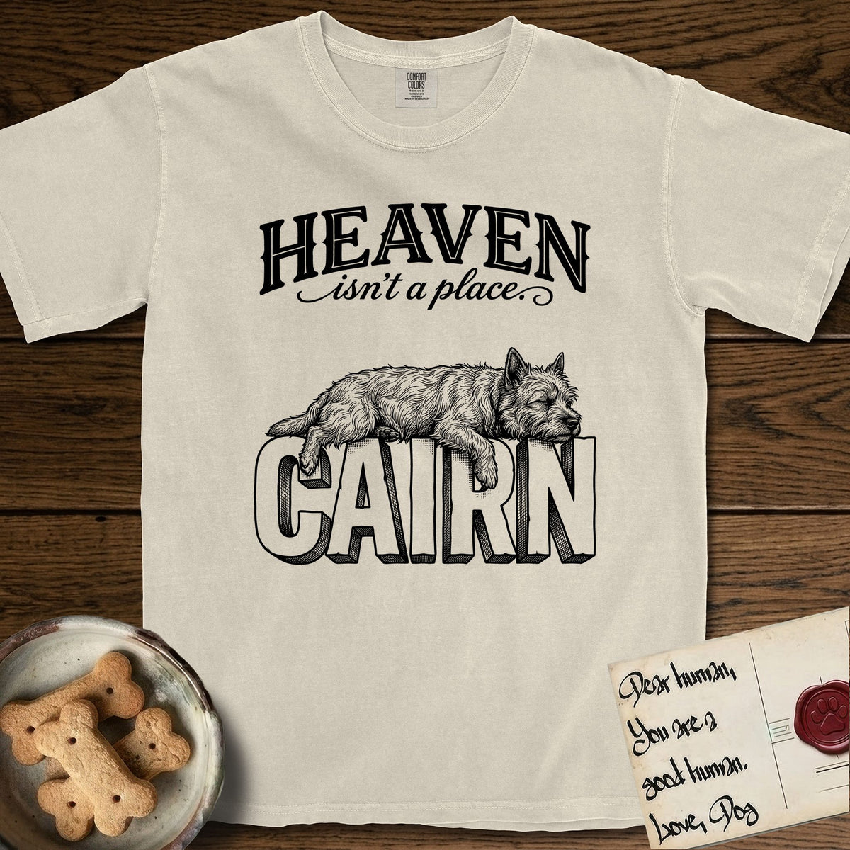 Heaven Isn't a Place Cairn Terrier T-Shirt flat lay in Ivory, featuring Black ink engraving illustration of a shaggy Cairn Terrier asleep on top of the giant carved block letters CAIRN, under the lettering HEAVEN isn't a place..