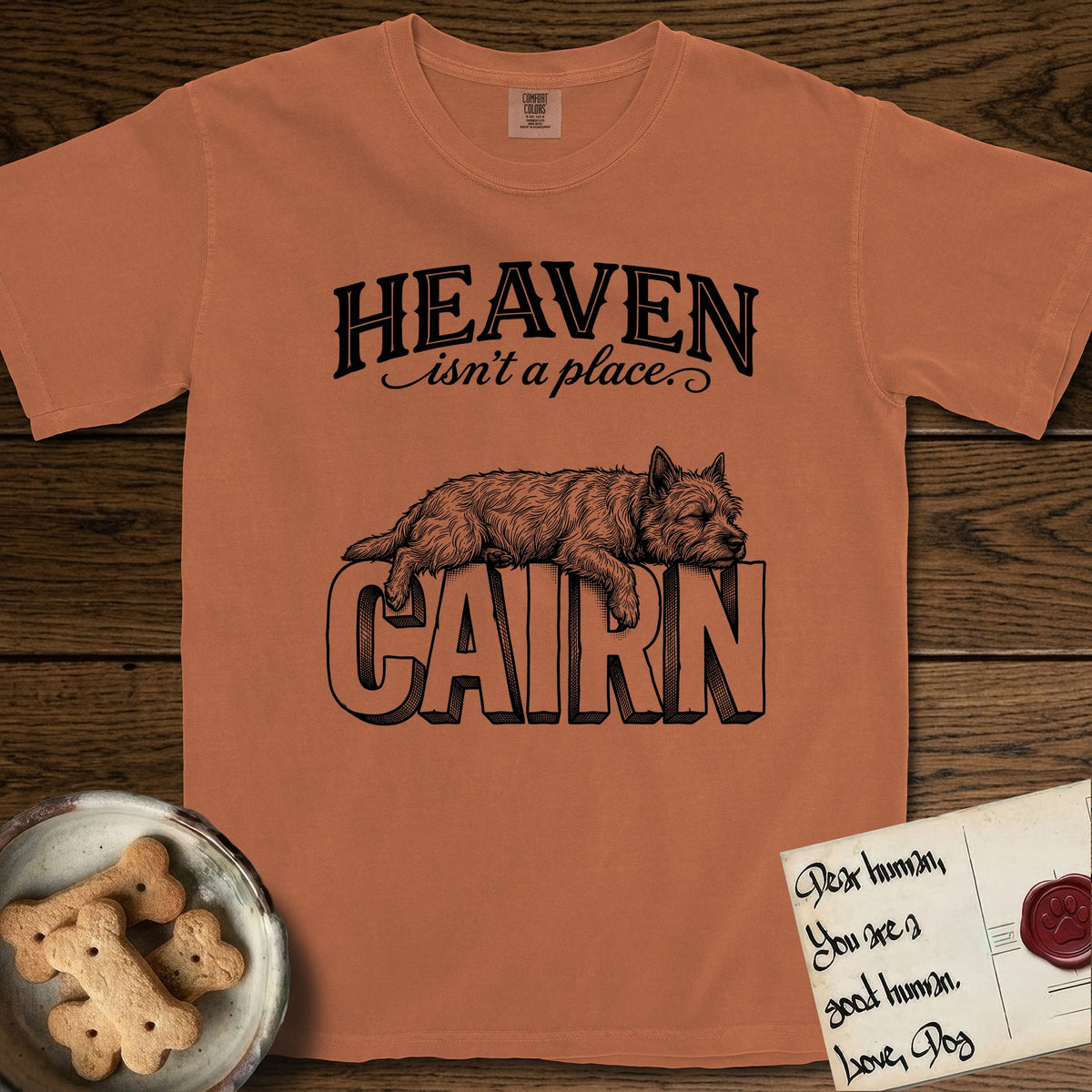 Heaven Isn't a Place Cairn Terrier T-Shirt flat lay in Yam, featuring Black ink engraving illustration of a shaggy Cairn Terrier asleep on top of the giant carved block letters CAIRN, under the lettering HEAVEN isn't a place..