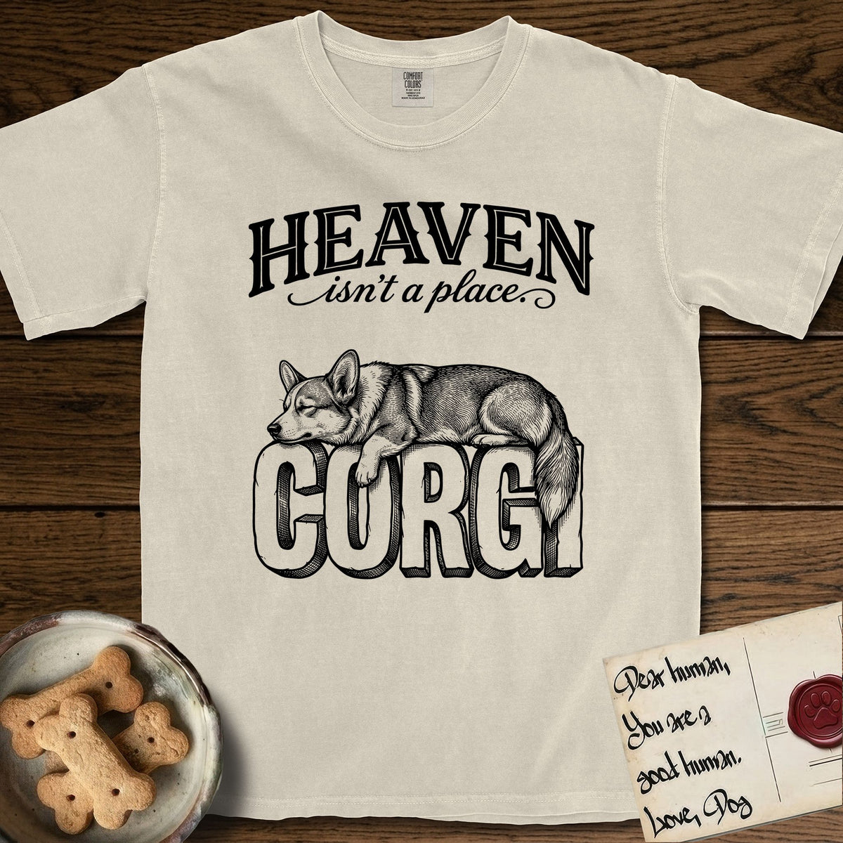 Heaven Isn't a Place Corgi T-Shirt flat lay in Ivory, featuring Black ink engraving illustration of a Corgi with a long fluffy tail asleep on top of the giant carved block letters CORGI, under the lettering HEAVEN isn't a place..