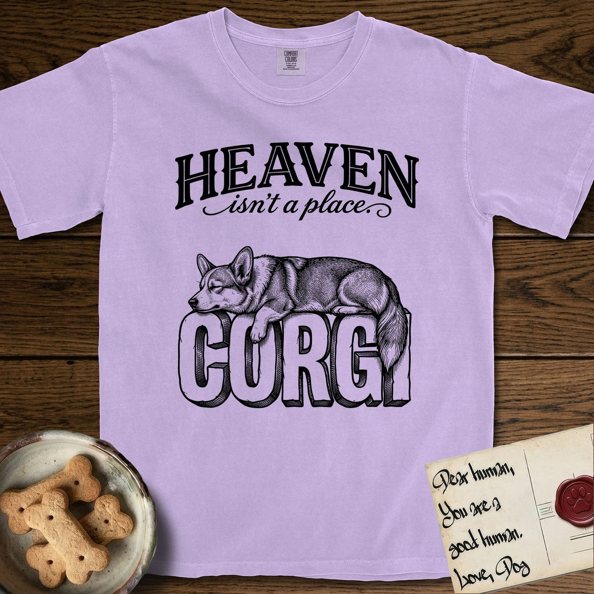 Heaven Isn't a Place Corgi T-Shirt flat lay in Orchid, featuring Black ink engraving illustration of a Corgi with a long fluffy tail asleep on top of the giant carved block letters CORGI, under the lettering HEAVEN isn't a place..