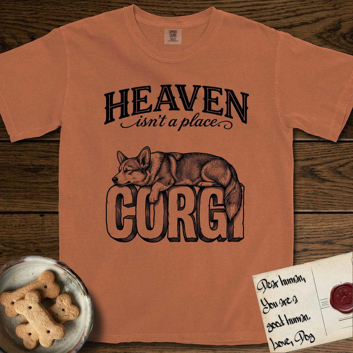 Heaven Isn't a Place Corgi T-Shirt flat lay in Yam, featuring Black ink engraving illustration of a Corgi with a long fluffy tail asleep on top of the giant carved block letters CORGI, under the lettering HEAVEN isn't a place..