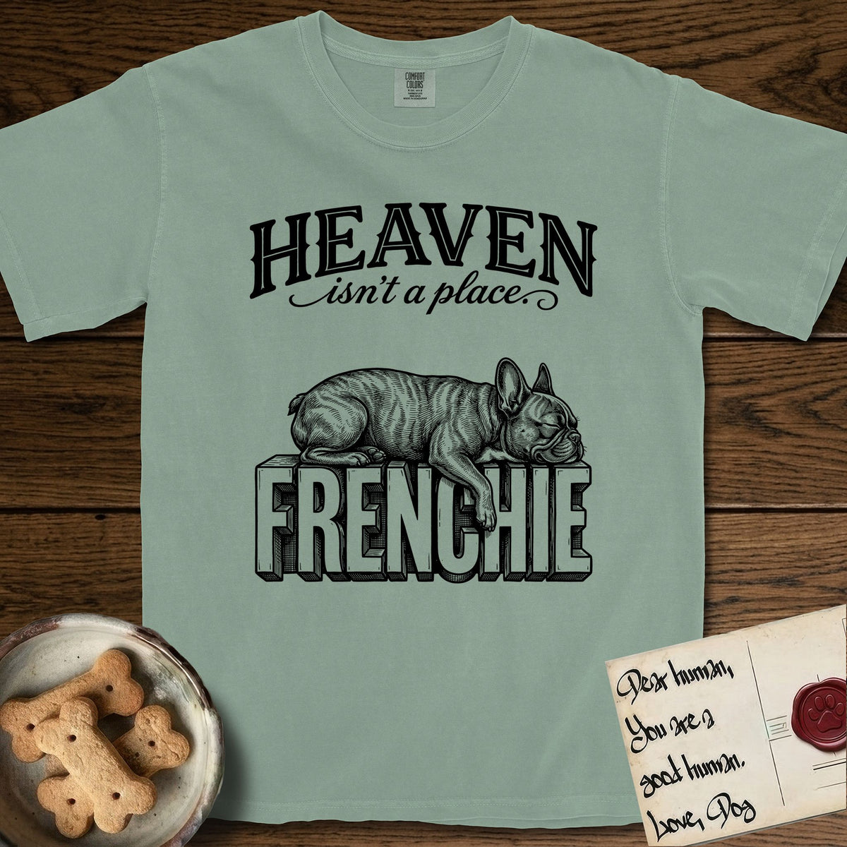 Heaven Isn't a Place French Bulldog T-Shirt flat lay in Bay, featuring Black ink engraving illustration of a brindle French Bulldog asleep on top of the giant carved block letters FRENCHIE, under the lettering HEAVEN isn't a place..
