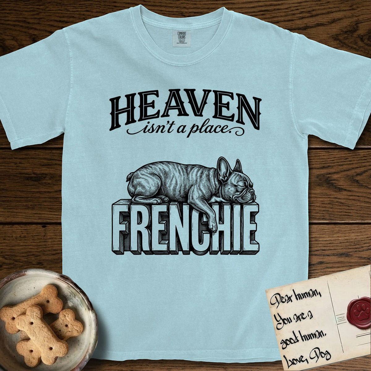 Heaven Isn't a Place French Bulldog T-Shirt flat lay in Chambray, featuring Black ink engraving illustration of a brindle French Bulldog asleep on top of the giant carved block letters FRENCHIE, under the lettering HEAVEN isn't a place..