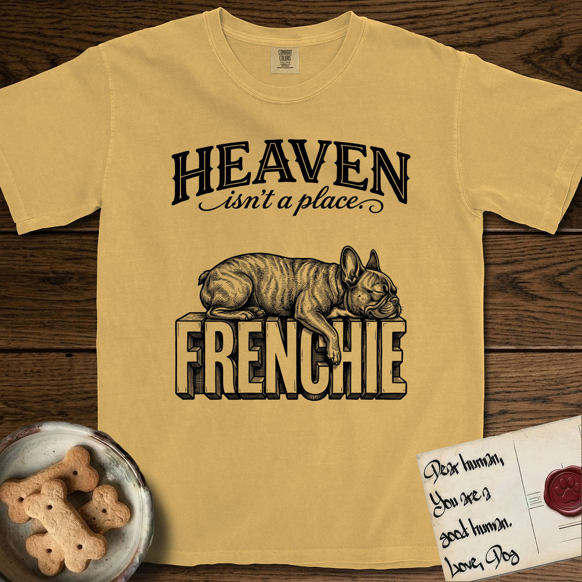 Heaven Isn't a Place French Bulldog T-Shirt flat lay in Mustard, featuring Black ink engraving illustration of a brindle French Bulldog asleep on top of the giant carved block letters FRENCHIE, under the lettering HEAVEN isn't a place..
