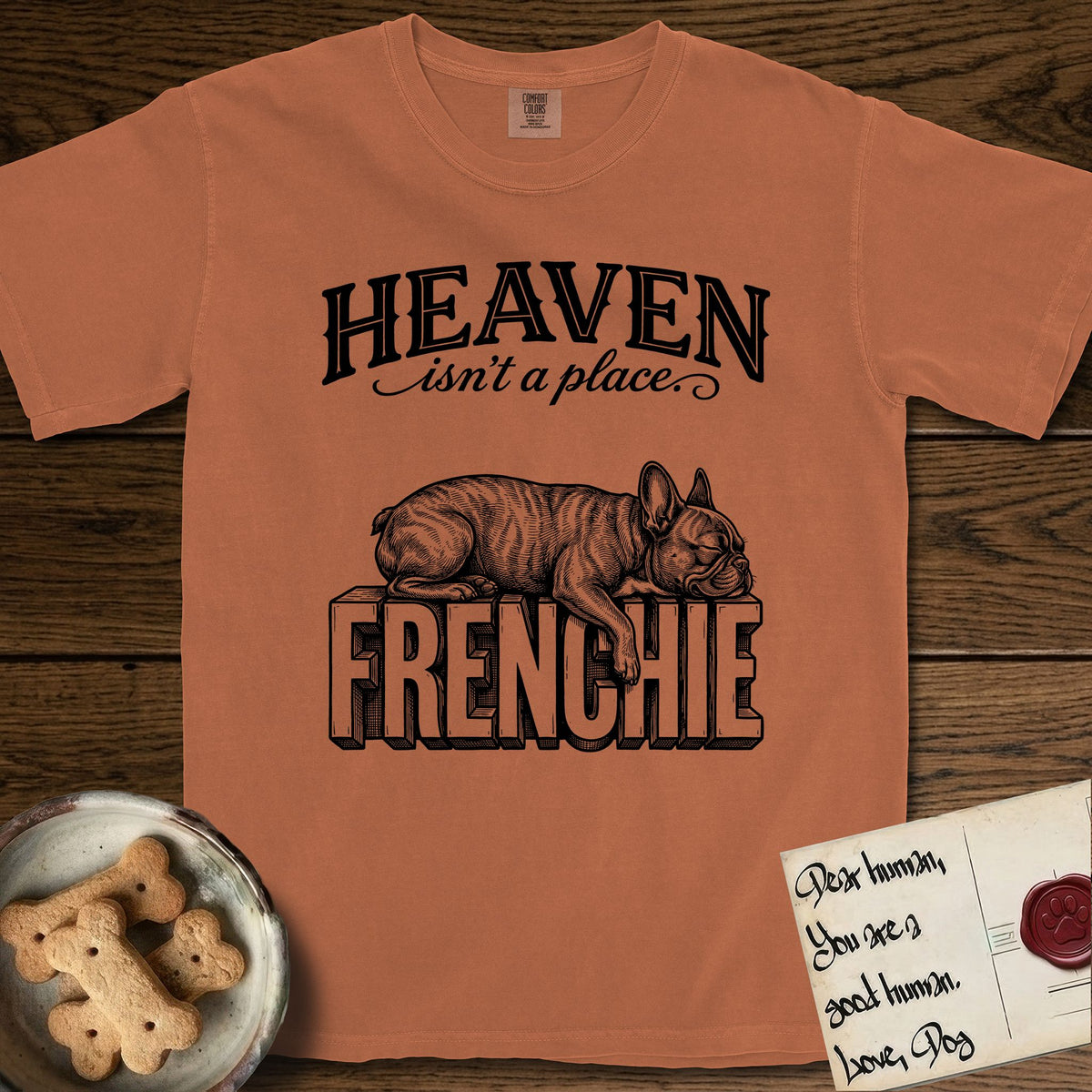 Heaven Isn't a Place French Bulldog T-Shirt flat lay in Yam, featuring Black ink engraving illustration of a brindle French Bulldog asleep on top of the giant carved block letters FRENCHIE, under the lettering HEAVEN isn't a place..