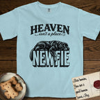 Heaven Isn't a Place Newfoundland T-Shirt flat lay in Chambray, featuring Black ink engraving illustration of a Newfoundland asleep on top of the giant carved block letters NEWFIE, under the lettering HEAVEN isn't a place..