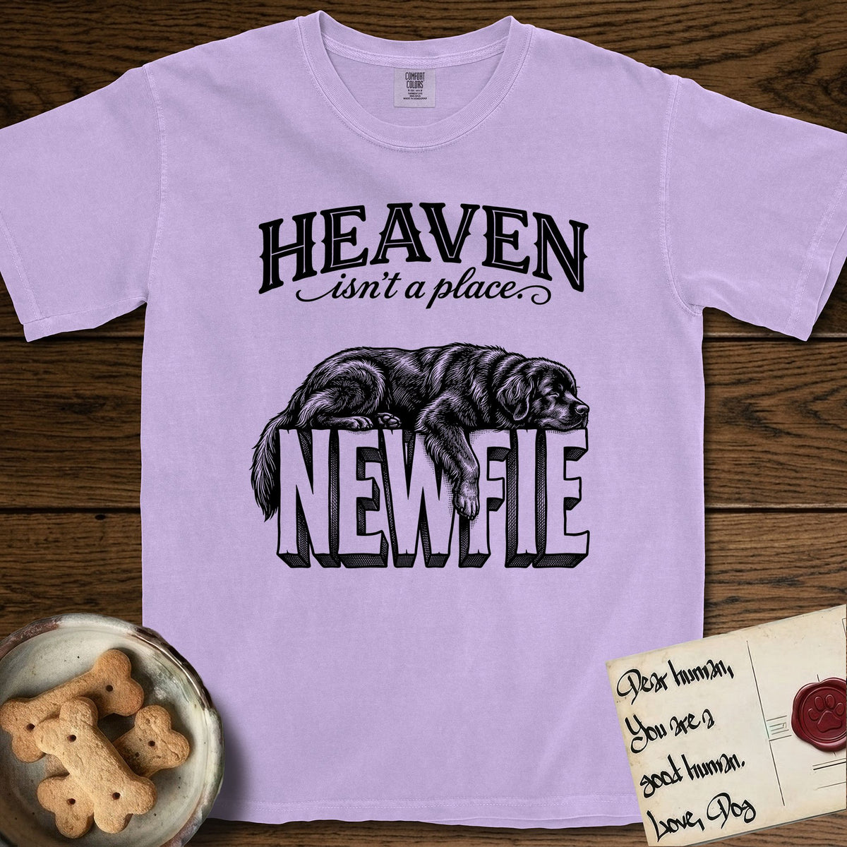 Heaven Isn't a Place Newfoundland T-Shirt flat lay in Orchid, featuring Black ink engraving illustration of a Newfoundland asleep on top of the giant carved block letters NEWFIE, under the lettering HEAVEN isn't a place..