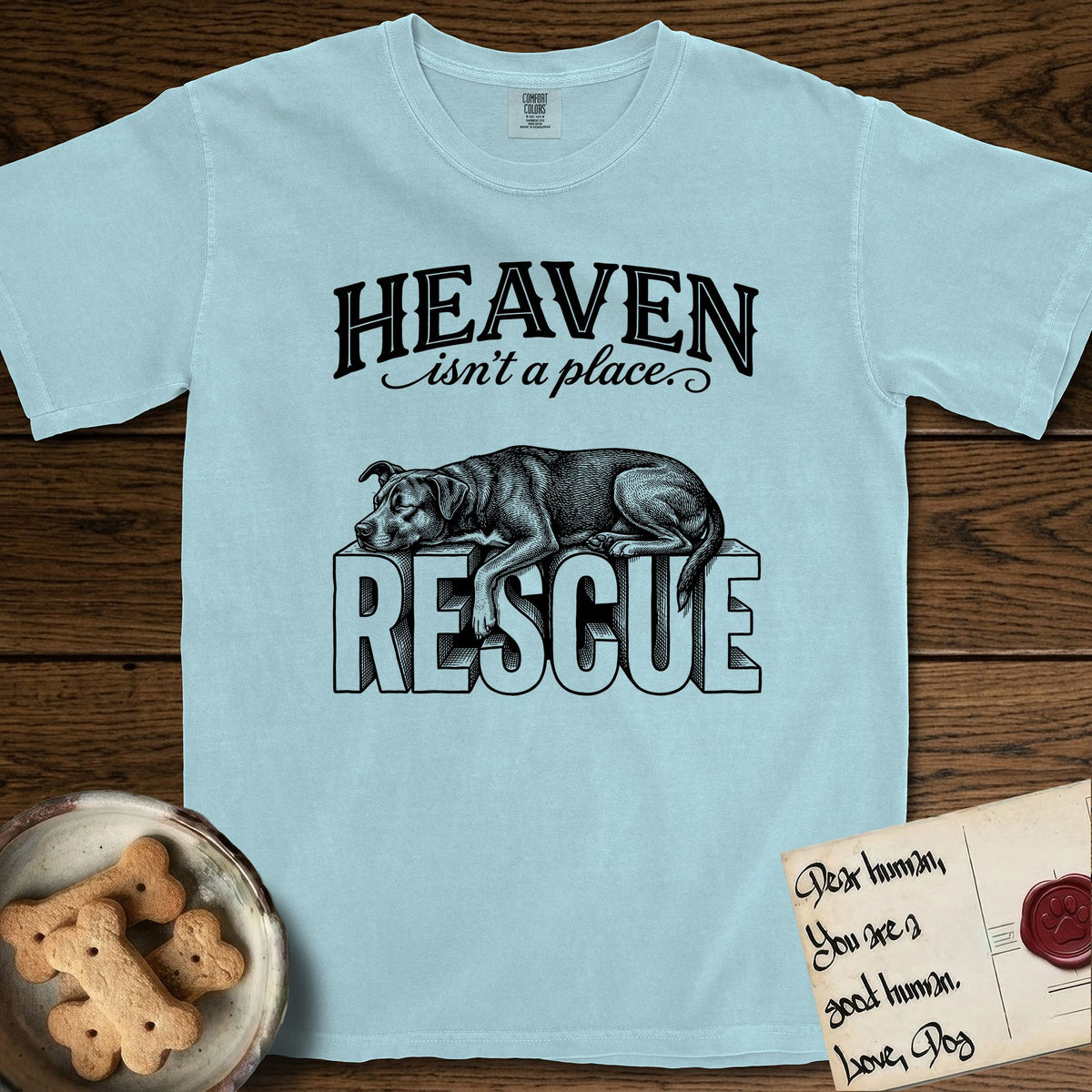 Heaven Isn't a Place Rescue Mutt T-Shirt flat lay in Chambray, featuring Black ink engraving illustration of a rescue Dog that is a Lab, Shepherd and Pitbull mix asleep on top of the giant carved block letters RESCUE, under the lettering HEAVEN isn't a place..