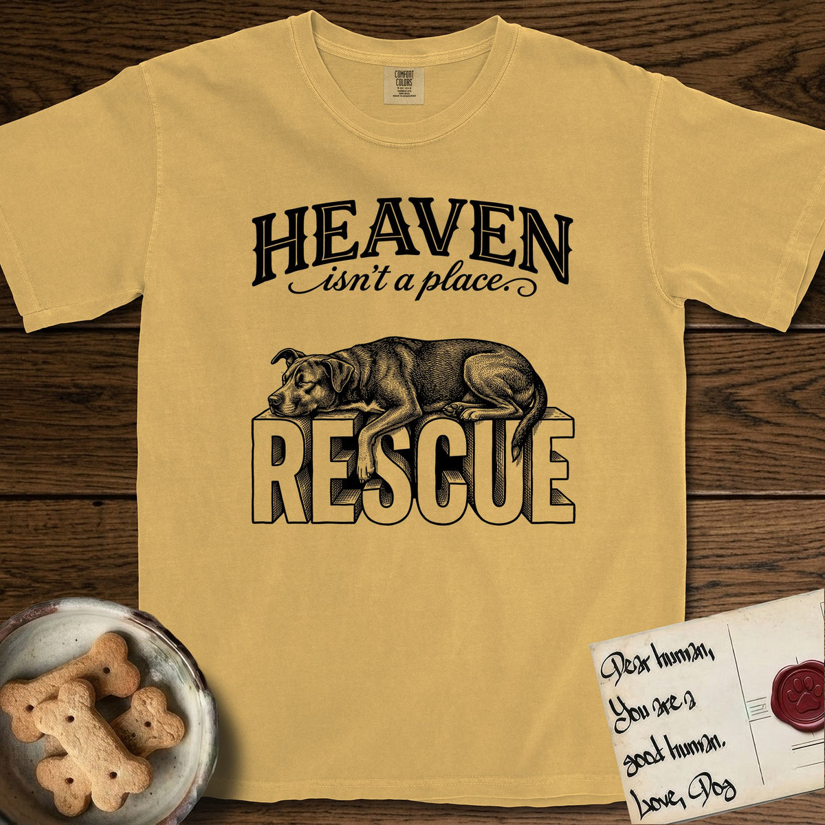 Heaven Isn't a Place Rescue Mutt T-Shirt flat lay in Mustard, featuring Black ink engraving illustration of a rescue Dog that is a Lab, Shepherd and Pitbull mix asleep on top of the giant carved block letters RESCUE, under the lettering HEAVEN isn't a place..