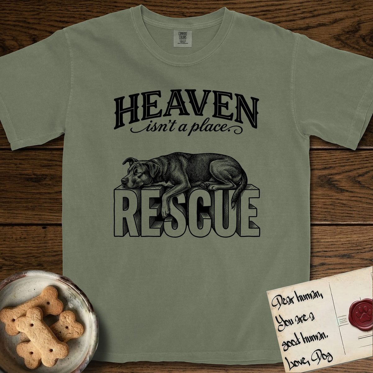 Heaven Isn't a Place Rescue Mutt T-Shirt flat lay in Sage, featuring Black ink engraving illustration of a rescue Dog that is a Lab, Shepherd and Pitbull mix asleep on top of the giant carved block letters RESCUE, under the lettering HEAVEN isn't a place..