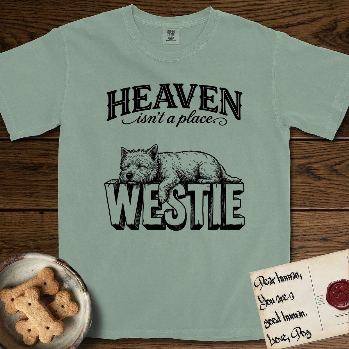 Heaven Isn't a Place Westie T-Shirt flat lay in Bay, featuring Black ink engraving illustration of a Westie asleep on top of the giant carved block letters WESTIE, under the lettering HEAVEN isn't a place..