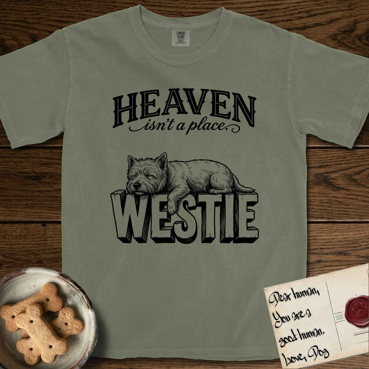 Heaven Isn't a Place Westie T-Shirt flat lay in Sage, featuring Black ink engraving illustration of a Westie asleep on top of the giant carved block letters WESTIE, under the lettering HEAVEN isn't a place..