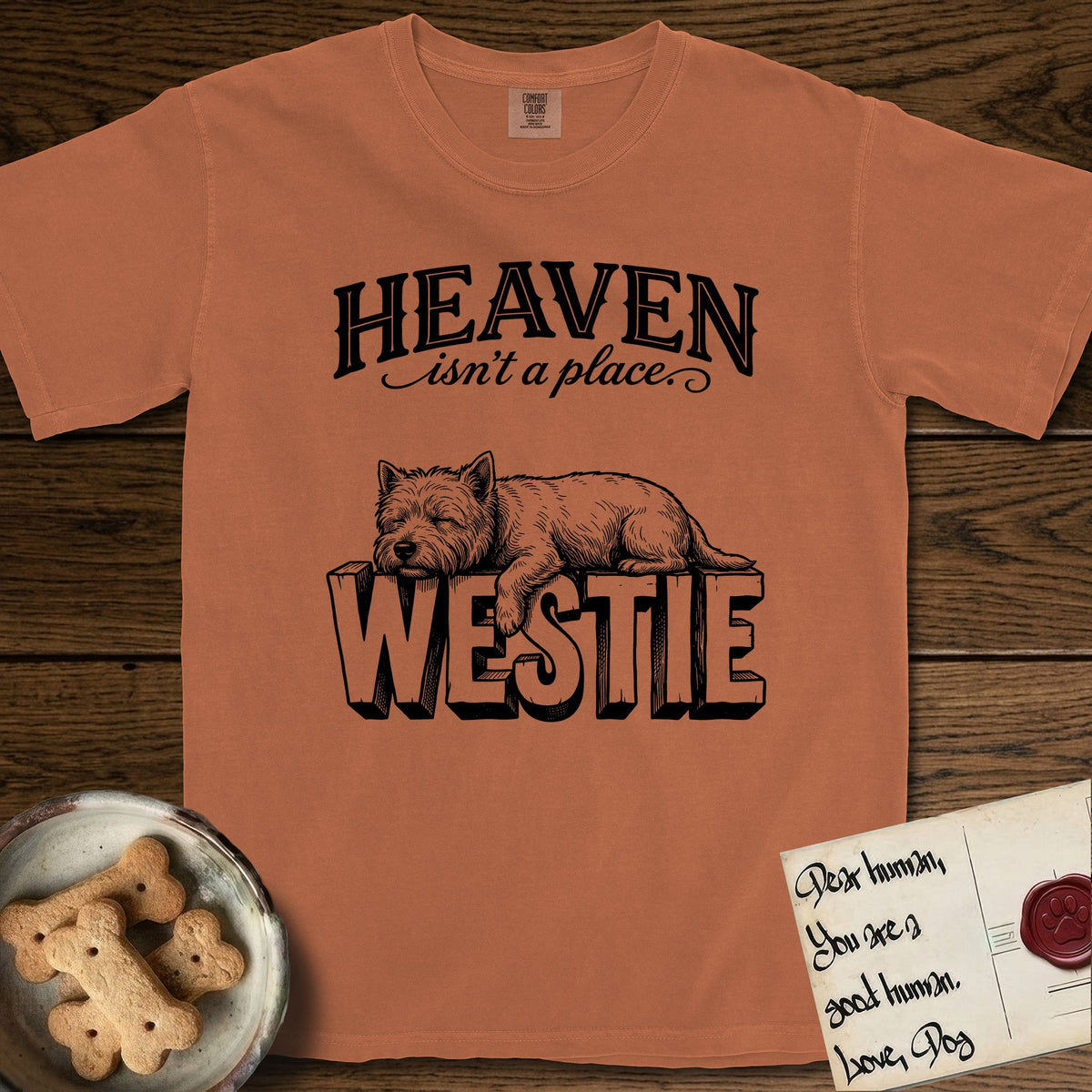 Heaven Isn't a Place Westie T-Shirt flat lay in Yam, featuring Black ink engraving illustration of a Westie asleep on top of the giant carved block letters WESTIE, under the lettering HEAVEN isn't a place..