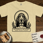 Holy Orders Basset Hound T-Shirt flat lay in Butter, featuring a bold black-and-white linocut saint-medallion portrait of a Basset Hound.