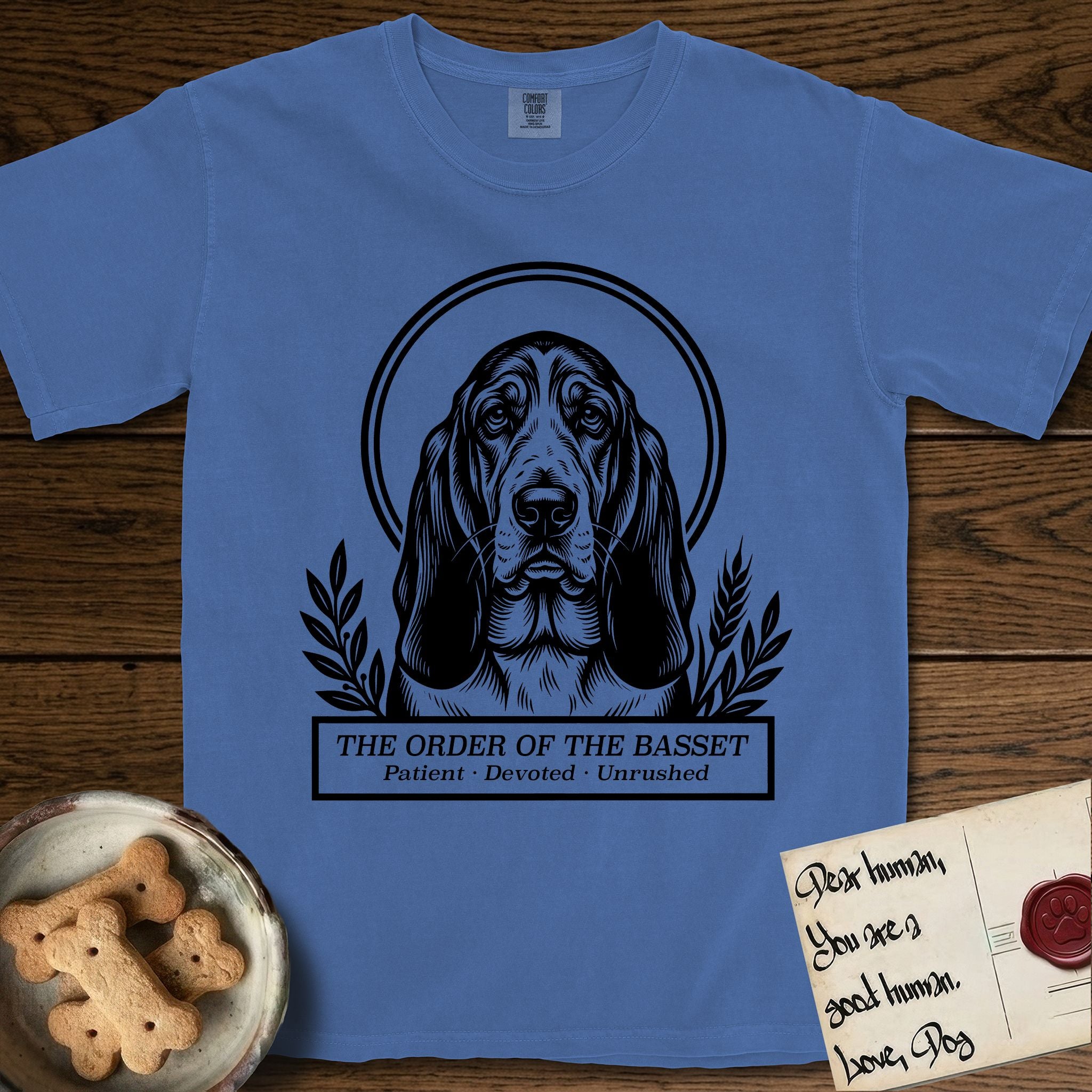 Holy Orders Basset Hound T-Shirt flat lay in Mystic Blue, featuring a bold black-and-white linocut saint-medallion portrait of a Basset Hound.