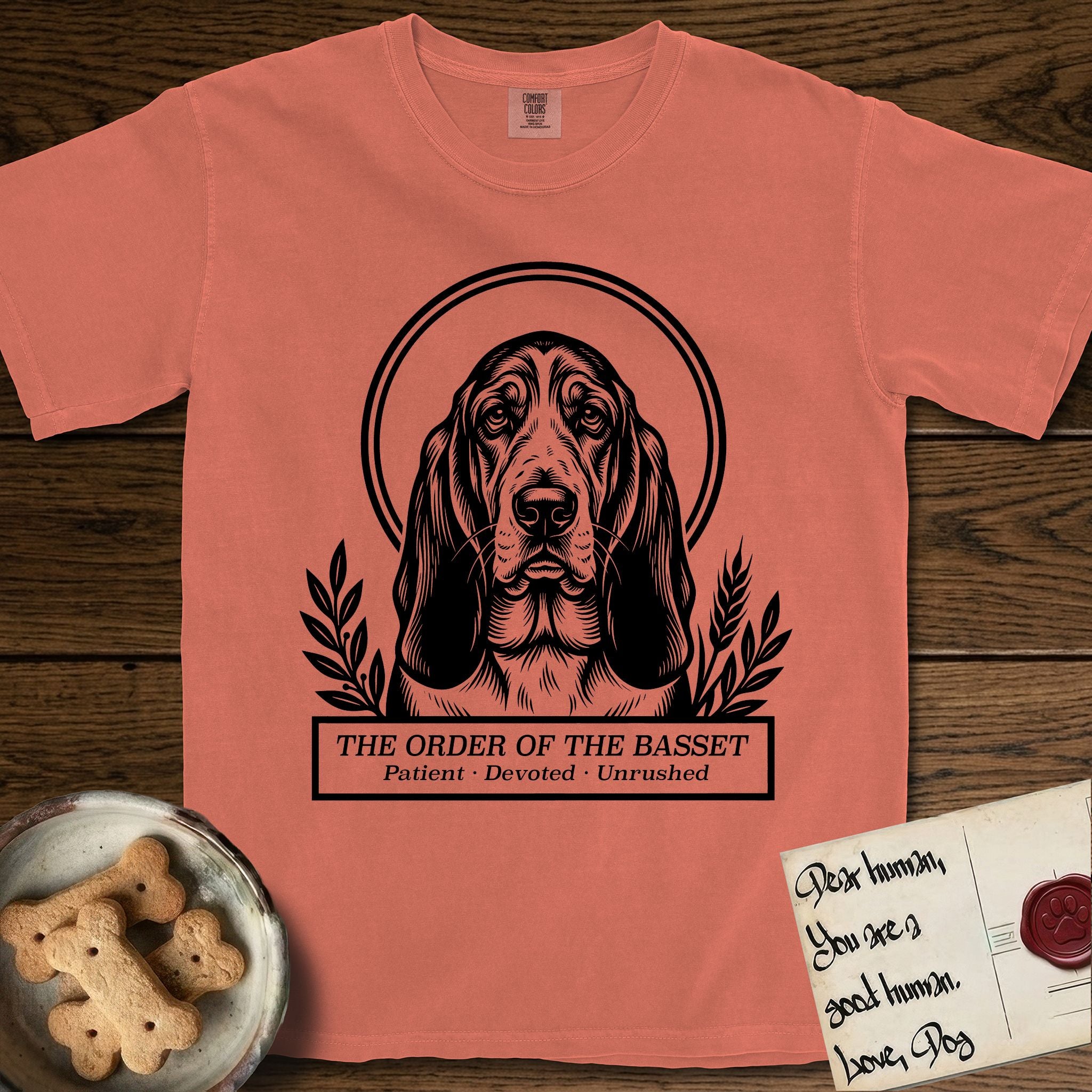 Holy Orders Basset Hound T-Shirt flat lay in Terracotta, featuring a bold black-and-white linocut saint-medallion portrait of a Basset Hound.