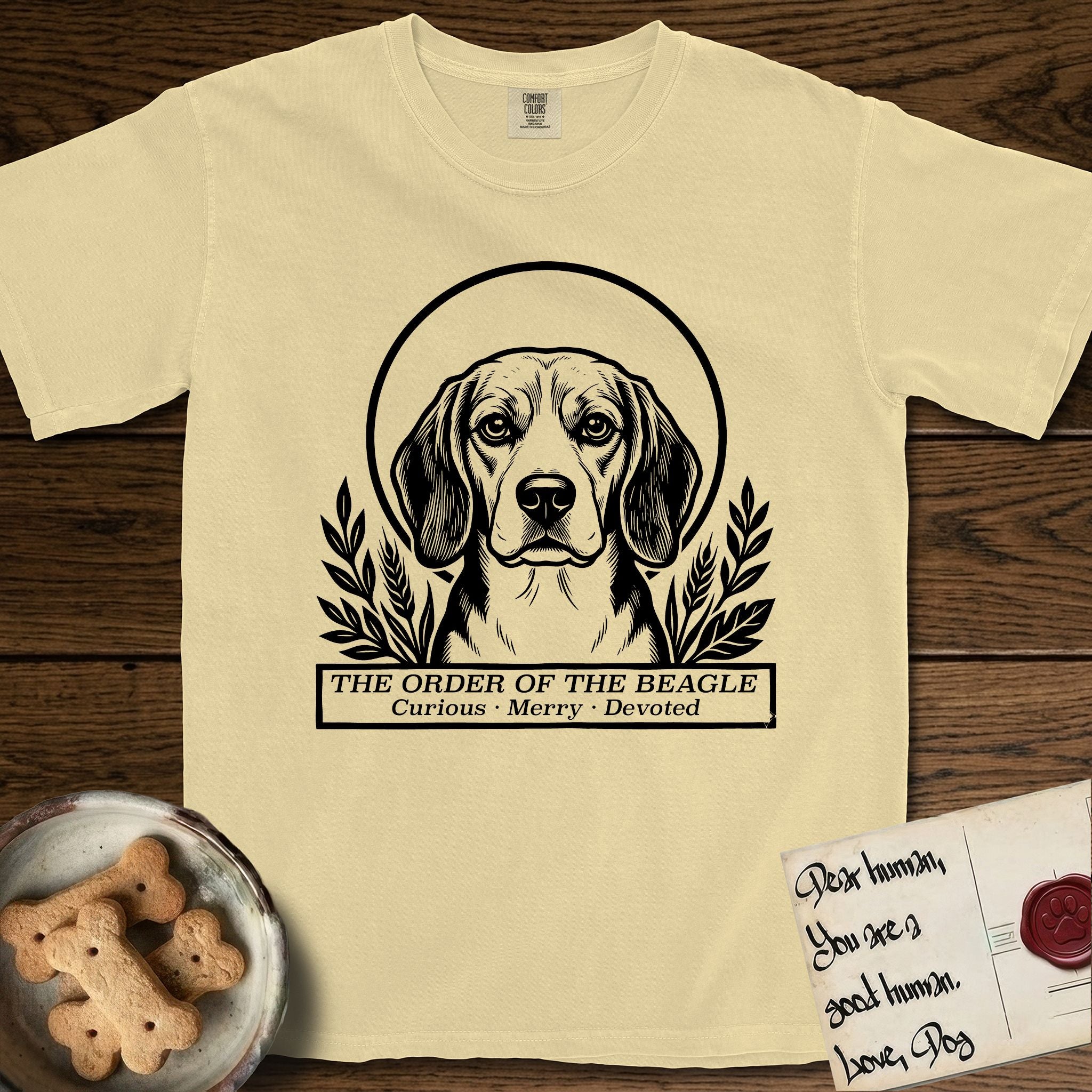 Holy Orders Beagle T-Shirt flat lay in Butter, featuring a bold black-and-white linocut saint-medallion portrait of a Beagle.