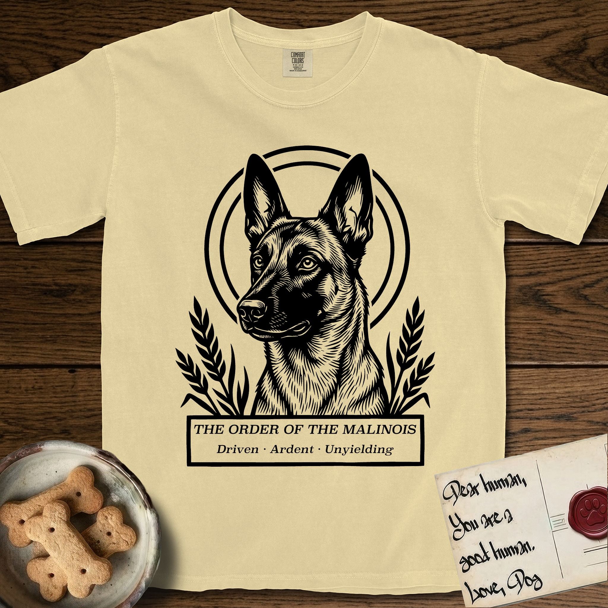 Holy Orders Belgian Malinois T-Shirt flat lay in Butter, featuring a bold black-and-white linocut saint-medallion portrait of a Belgian Malinois.