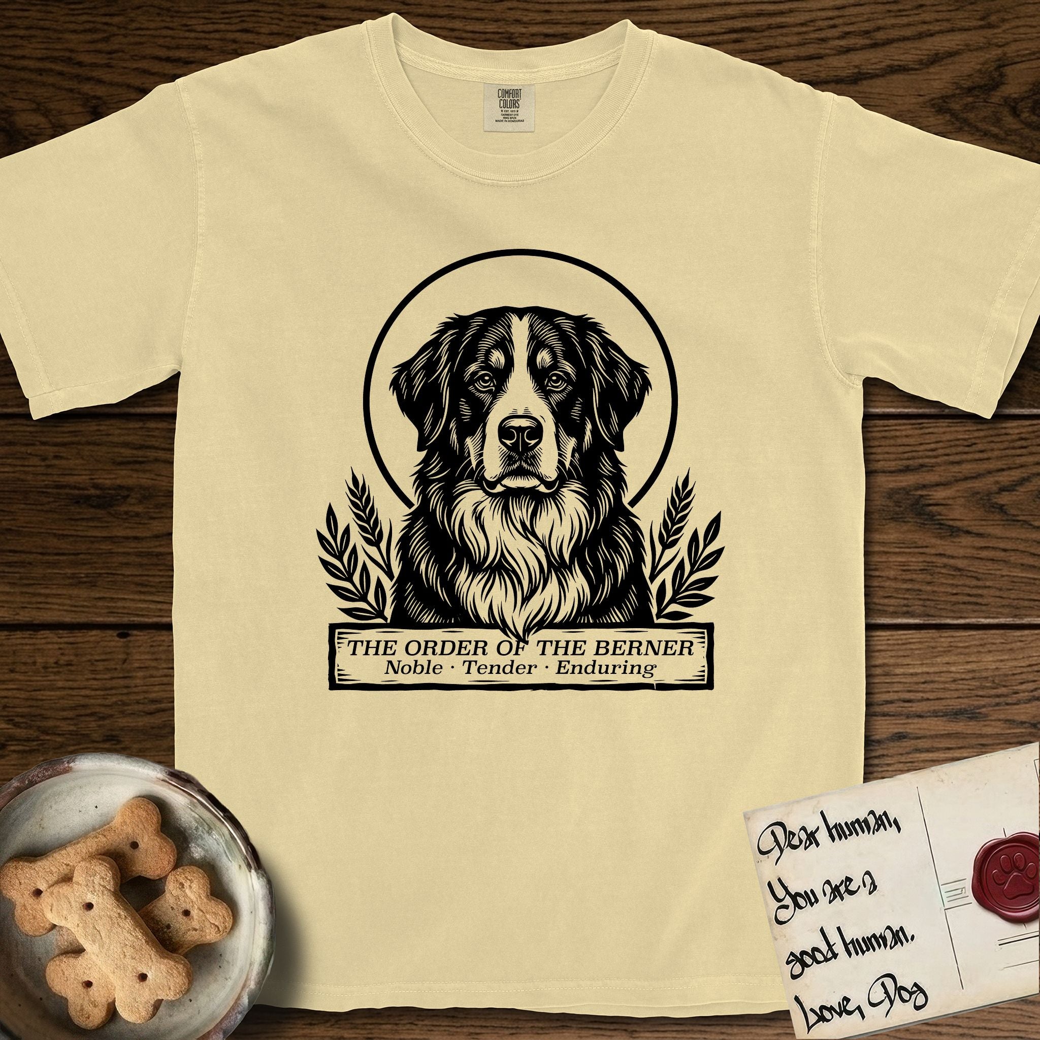 Holy Orders Bernese Mountain Dog T-Shirt flat lay in Butter, featuring a bold black-and-white linocut saint-medallion portrait of a Bernese Mountain Dog.