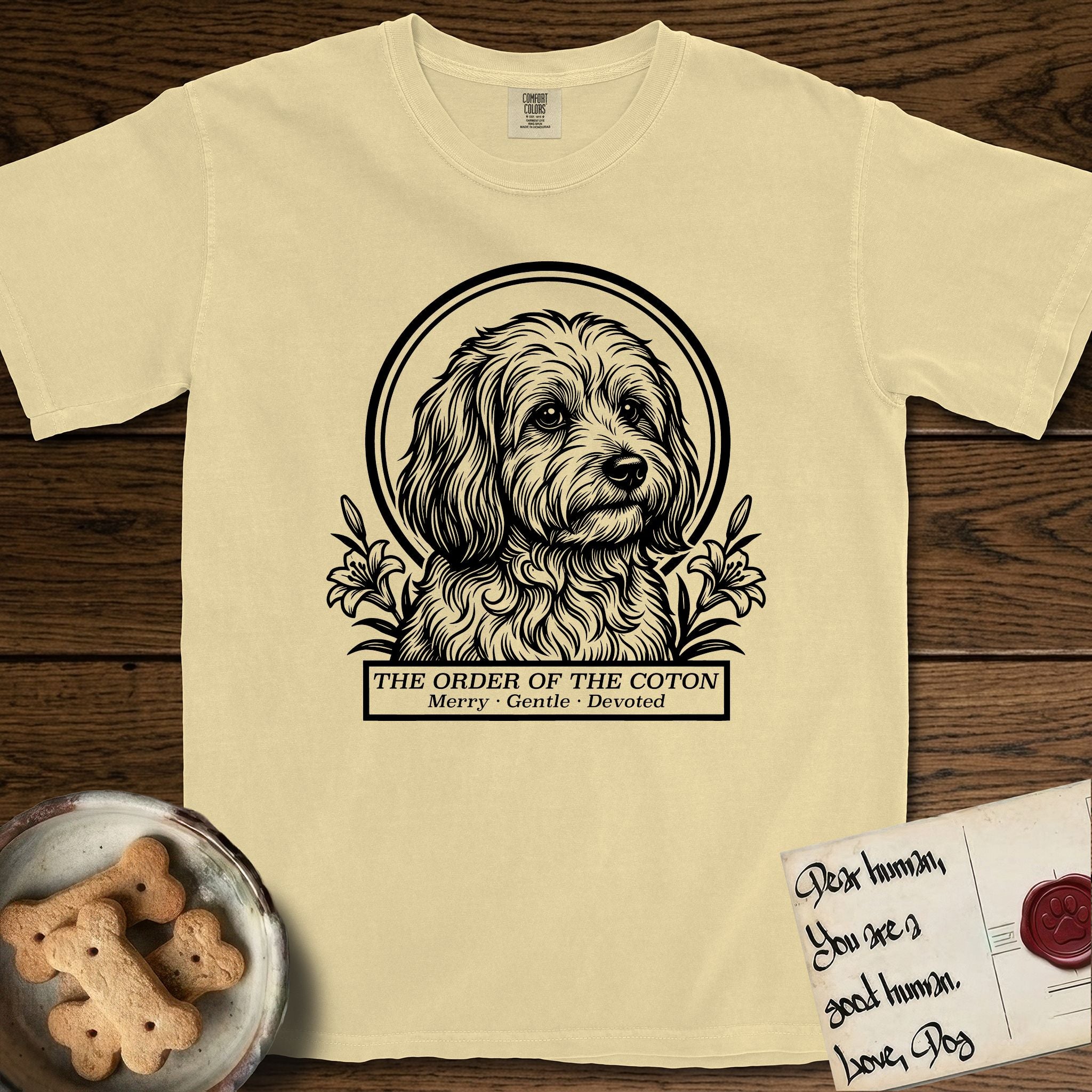 Holy Orders Coton T-Shirt flat lay in Butter, featuring a bold black-and-white linocut saint-medallion portrait of a Coton de Tulear.