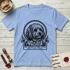 Holy Orders Coton T-Shirt flat lay in Light Blue, featuring a bold black-and-white linocut saint-medallion portrait of a Coton de Tulear.