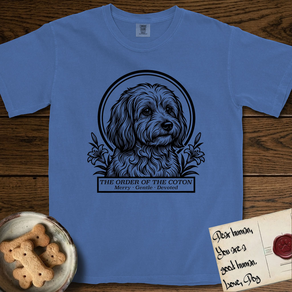 Holy Orders Coton T-Shirt flat lay in Mystic Blue, featuring a bold black-and-white linocut saint-medallion portrait of a Coton de Tulear.