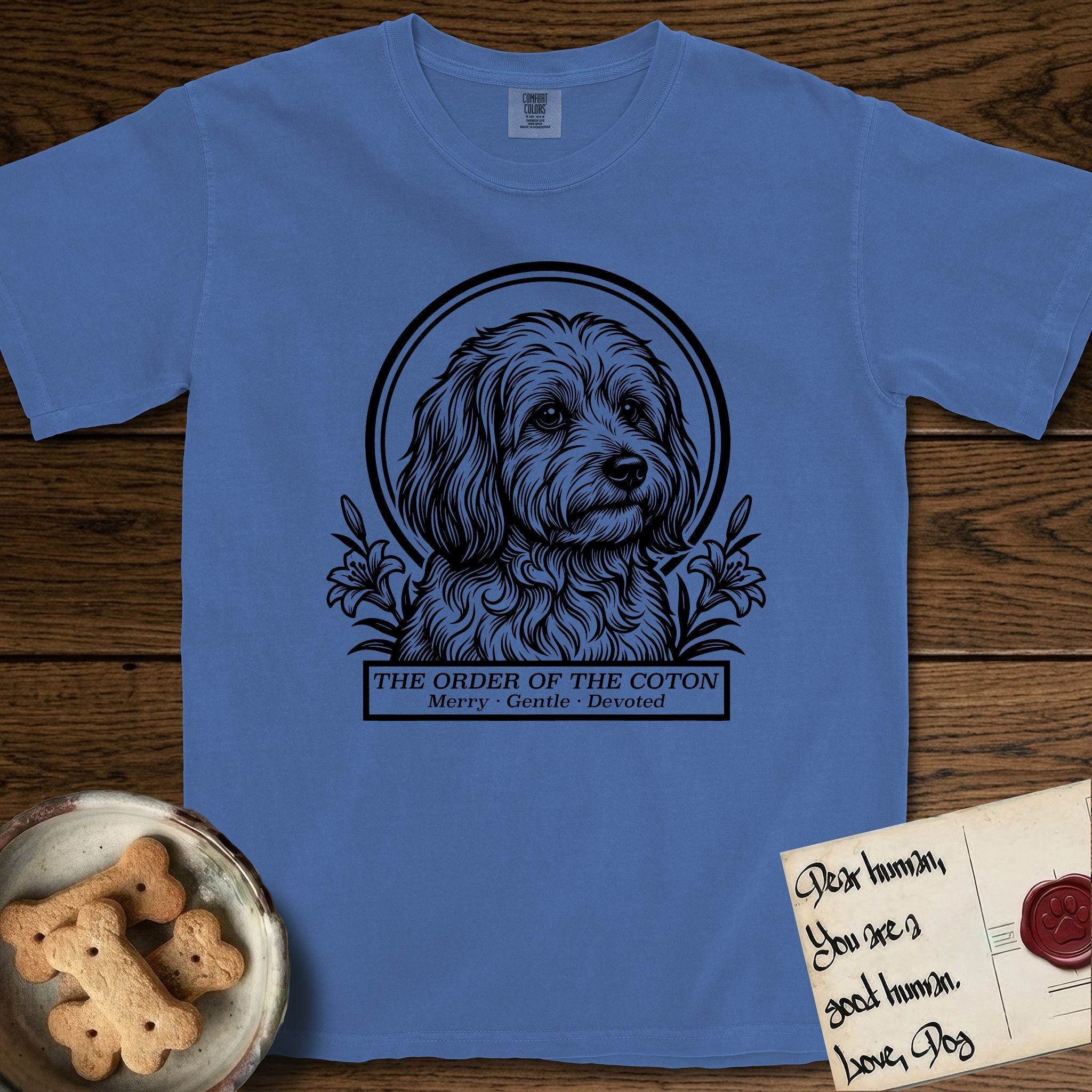 Holy Orders Coton T-Shirt flat lay in Mystic Blue, featuring a bold black-and-white linocut saint-medallion portrait of a Coton de Tulear.