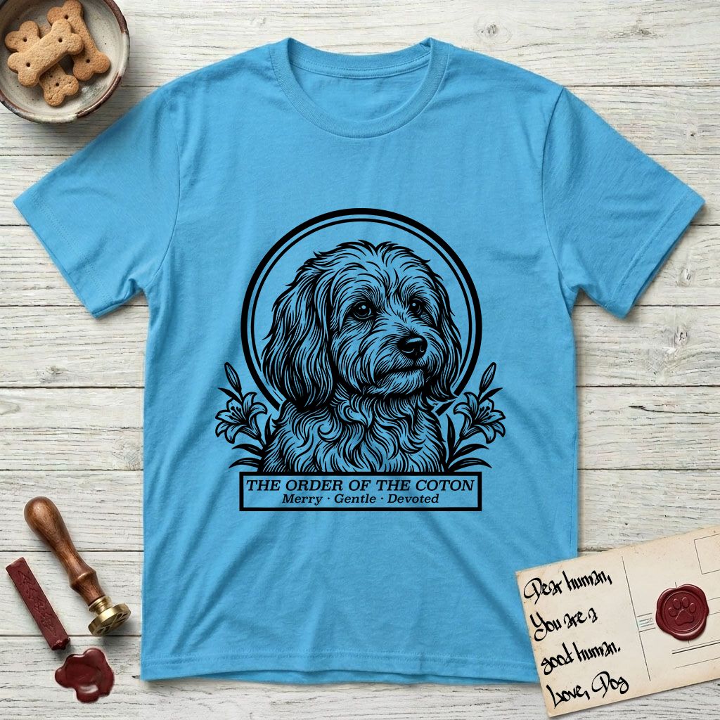 Holy Orders Coton T-Shirt flat lay in Sky, featuring a bold black-and-white linocut saint-medallion portrait of a Coton de Tulear.