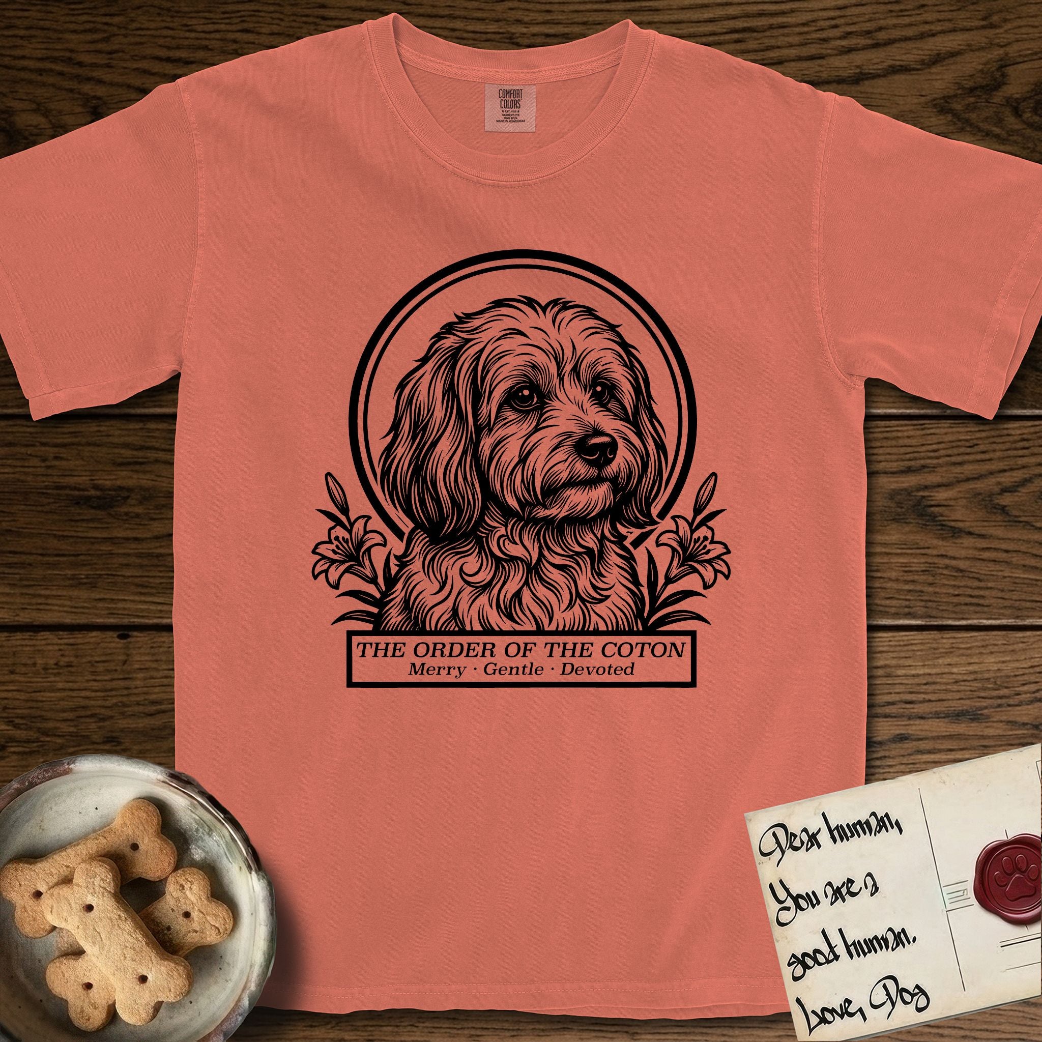 Holy Orders Coton T-Shirt flat lay in Terracotta, featuring a bold black-and-white linocut saint-medallion portrait of a Coton de Tulear.
