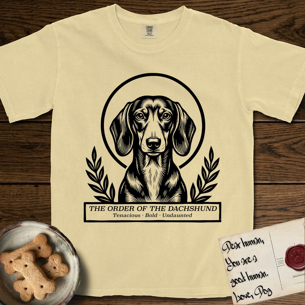 Holy Orders Dachshund T-Shirt flat lay in Butter, featuring a bold black-and-white linocut saint-medallion portrait of a Dachshund.