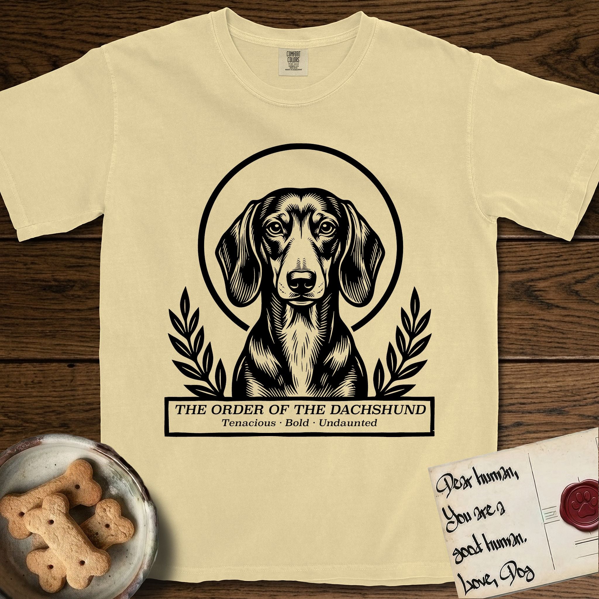 Holy Orders Dachshund T-Shirt flat lay in Butter, featuring a bold black-and-white linocut saint-medallion portrait of a Dachshund.