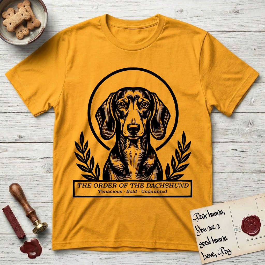 Holy Orders Dachshund T-Shirt flat lay in Gold, featuring a bold black-and-white linocut saint-medallion portrait of a Dachshund.