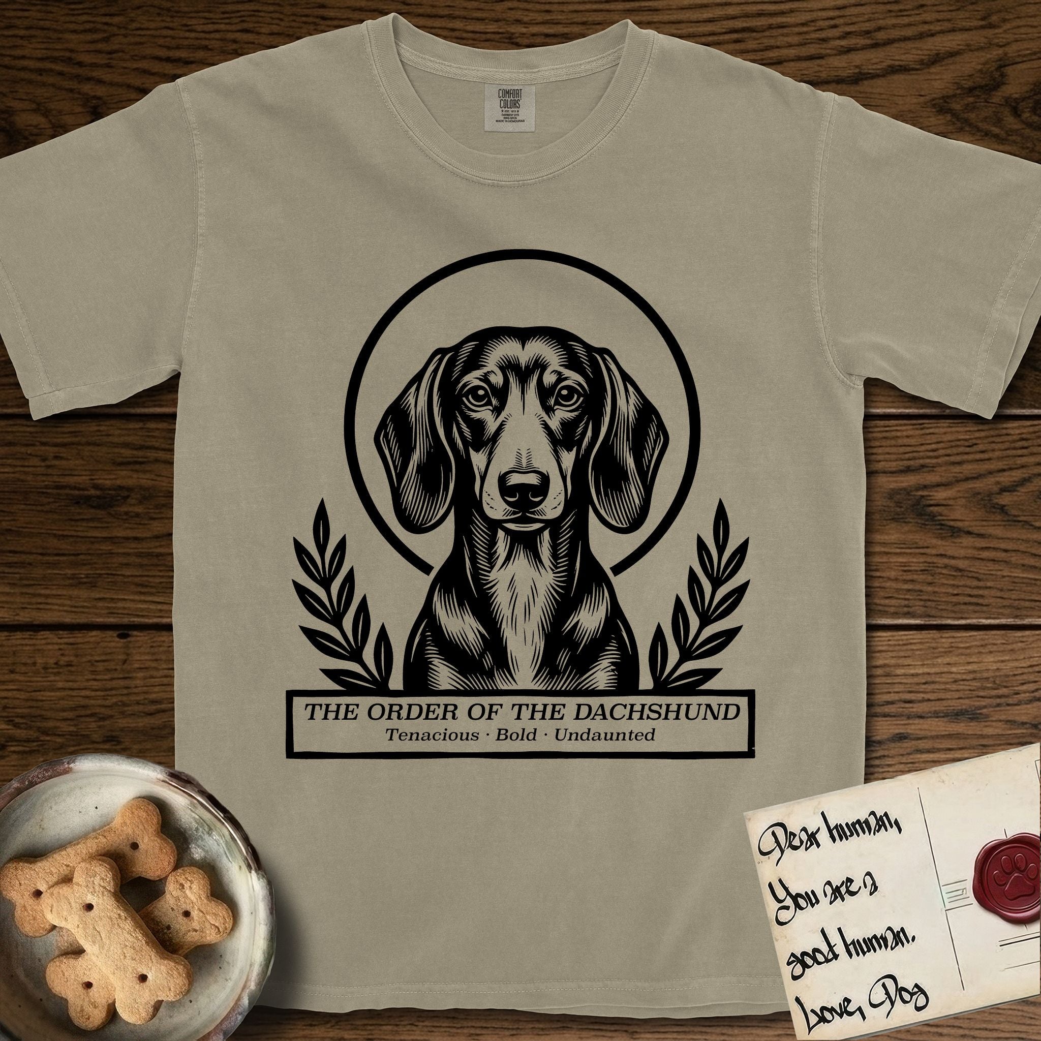 Holy Orders Dachshund T-Shirt flat lay in Khaki, featuring a bold black-and-white linocut saint-medallion portrait of a Dachshund.