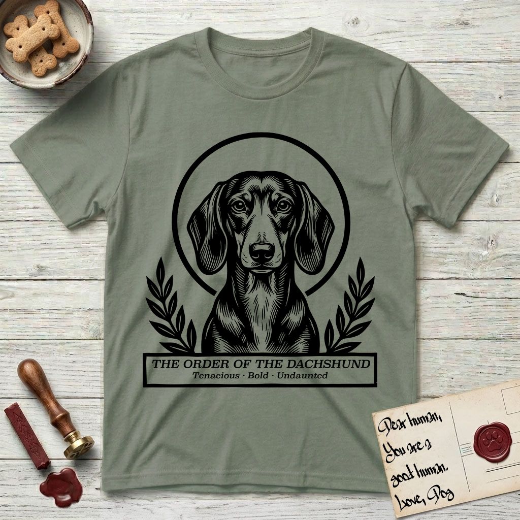 Holy Orders Dachshund T-Shirt flat lay in Sage, featuring a bold black-and-white linocut saint-medallion portrait of a Dachshund.
