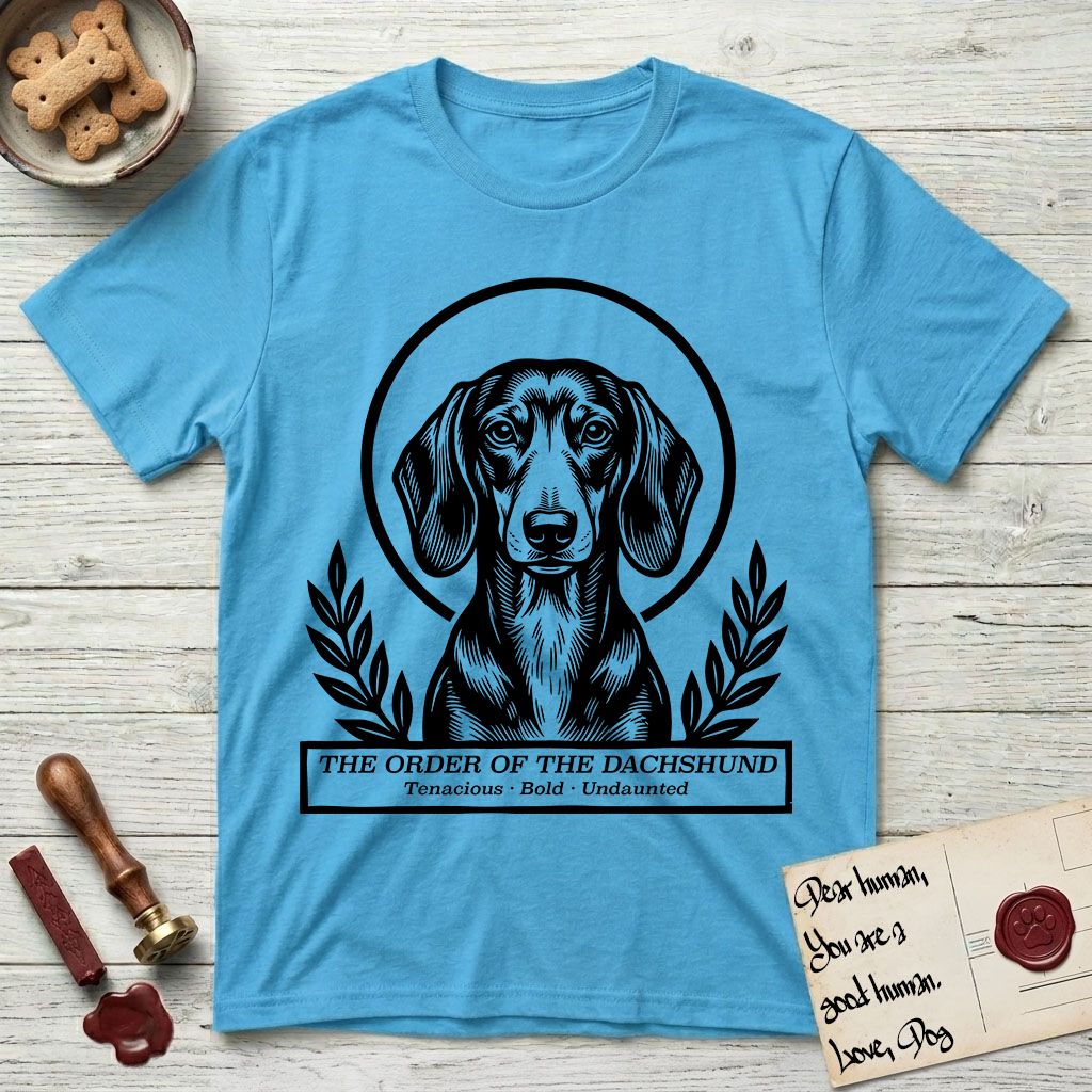 Holy Orders Dachshund T-Shirt flat lay in Sky, featuring a bold black-and-white linocut saint-medallion portrait of a Dachshund.