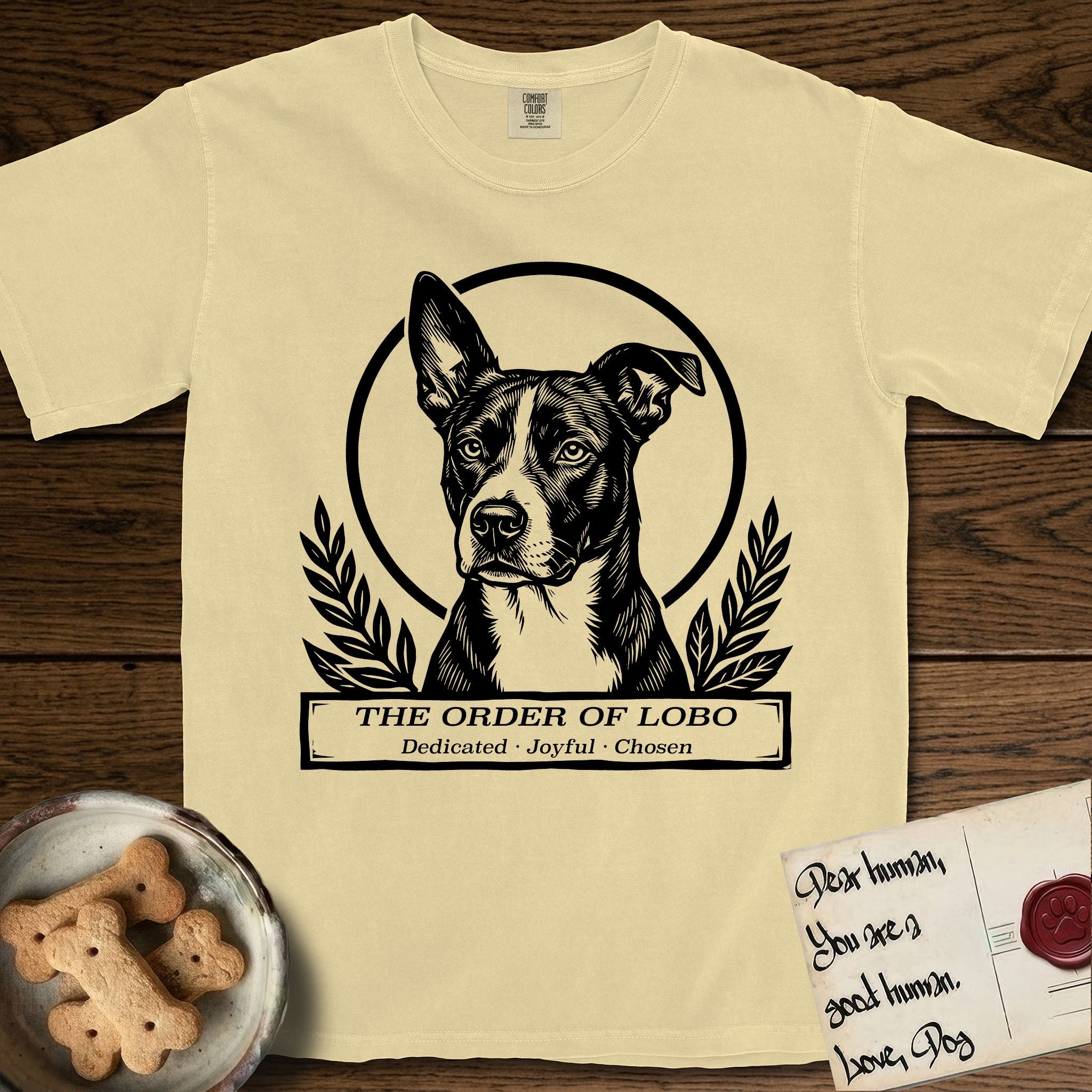 Holy Orders Lobo T-Shirt flat lay in Butter, featuring a bold black-and-white linocut saint-medallion portrait of Lobo, an Australian Cattle Dog and Pitbull mix with a white facial blaze.