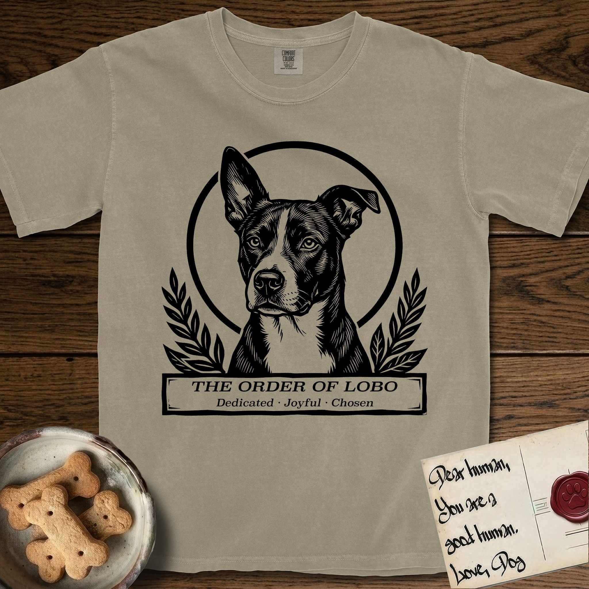 Holy Orders Lobo T-Shirt flat lay in Khaki, featuring a bold black-and-white linocut saint-medallion portrait of Lobo, an Australian Cattle Dog and Pitbull mix with a white facial blaze.