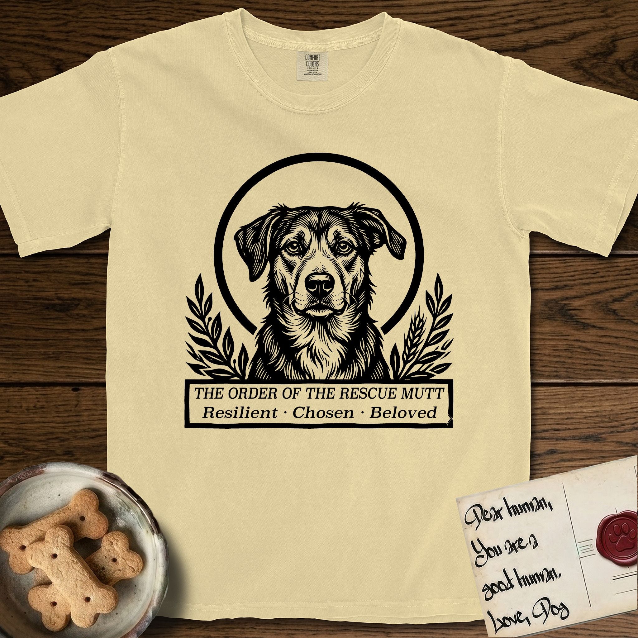 Holy Orders Rescue Mutt T-Shirt flat lay in Butter, featuring a bold black-and-white linocut saint-medallion portrait of a rescue mutt.