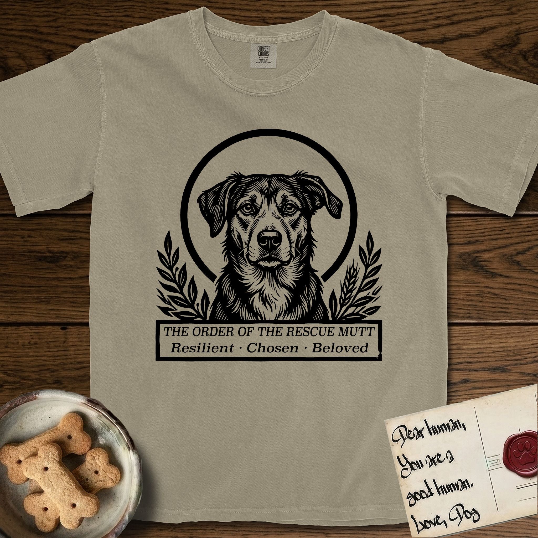 Holy Orders Rescue Mutt T-Shirt flat lay in Khaki, featuring a bold black-and-white linocut saint-medallion portrait of a rescue mutt.