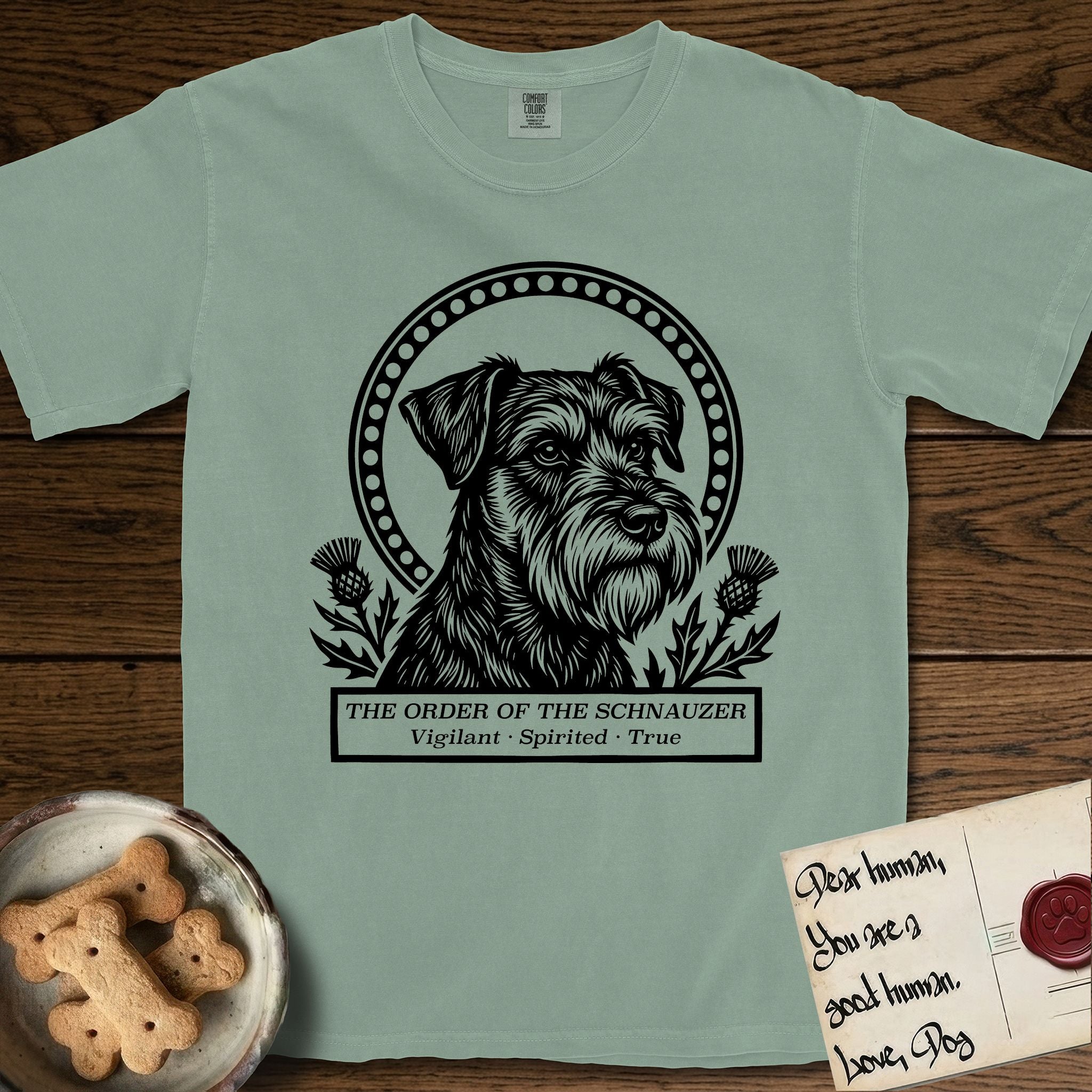Holy Orders Schnauzer T-Shirt flat lay in Bay, featuring a bold black-and-white linocut saint-medallion portrait of a Schnauzer.
